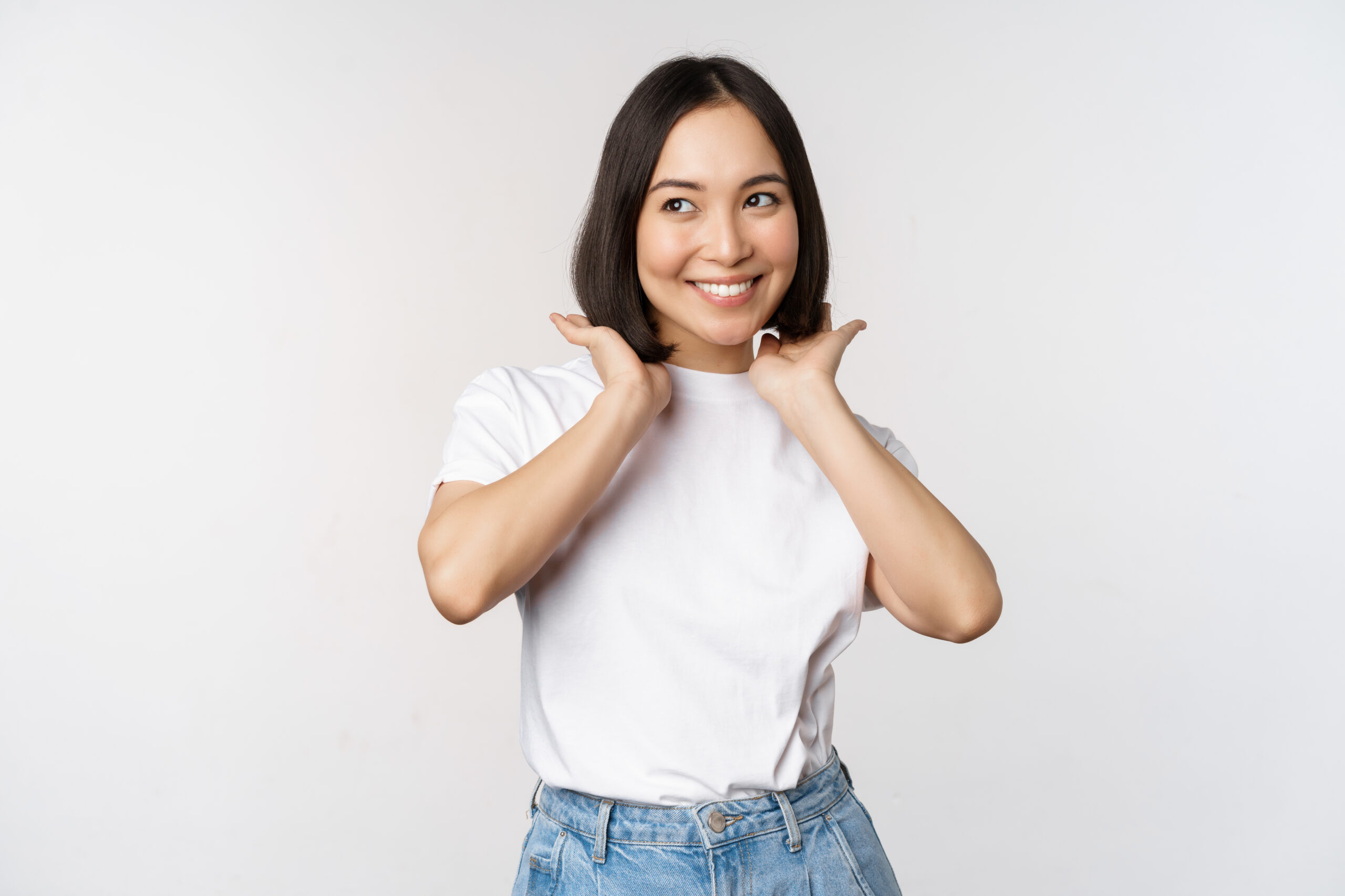 portrait of cute, beautiful asian woman touching her new short haircut, showing hairstyle, smiling happy at camera, standing over white background